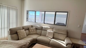 A corner living room with a beige sectional sofa, a small wooden table, and a large window with blinds. - Cool & Calm - walk to the beach (SOUTH WEST ROCKS)