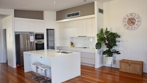 A modern kitchen with white cabinetry, a central island with bar stools, a built-in oven, and a large clock on the wall. - Cool & Calm - walk to the beach (SOUTH WEST ROCKS)