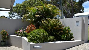 A landscaped garden area with various plants and shrubs, a concrete planter, and a white wall. - Cool & Calm - walk to the beach (SOUTH WEST ROCKS)