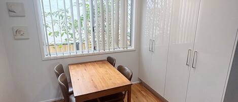 Um quarto com mesa de madeira, cadeiras, janela com persianas e armários brancos.