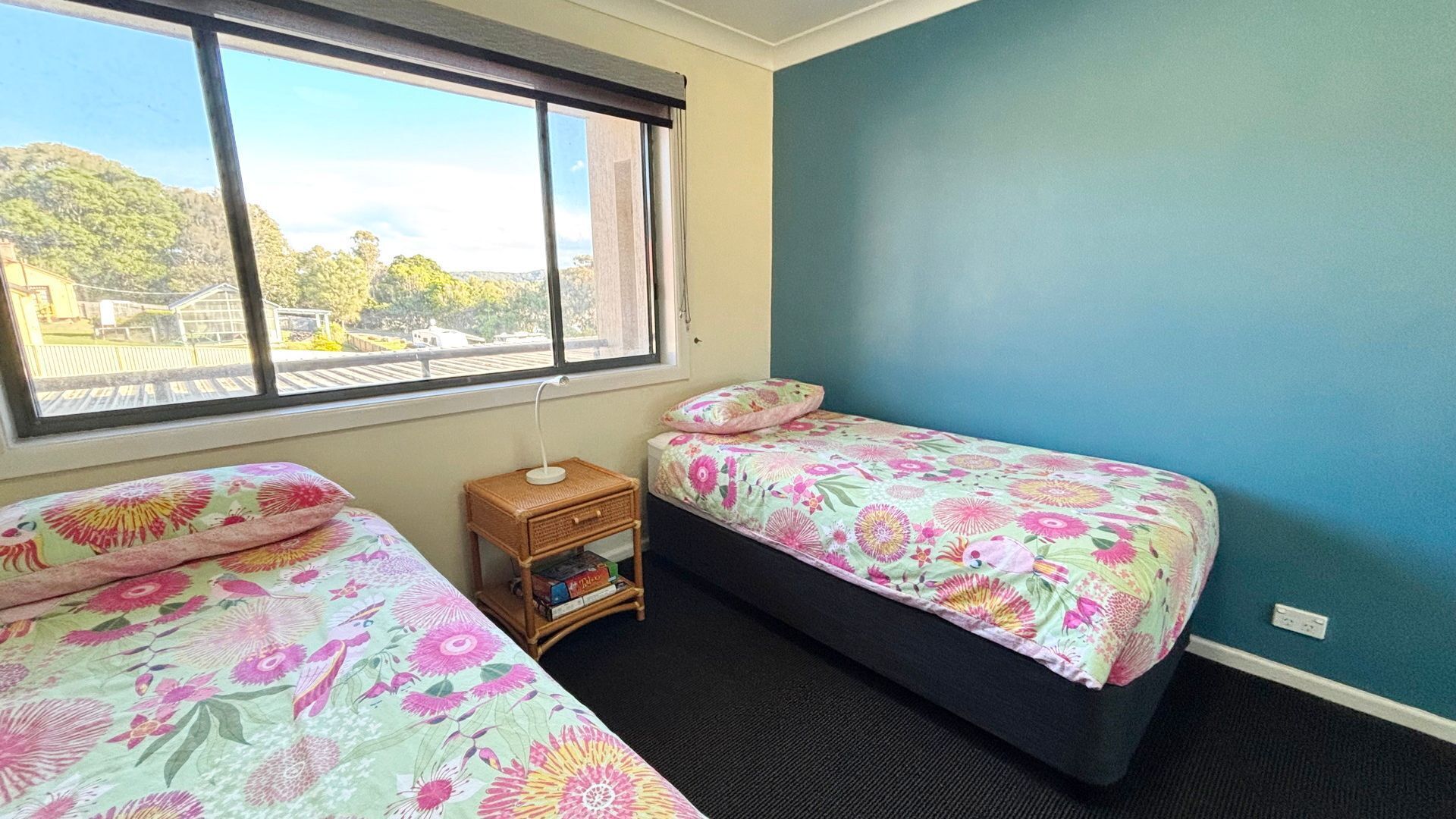 A bedroom with two beds, a small bedside table, a window, and a view of greenery outside.