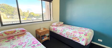 A bedroom with two beds, a small bedside table, a window, and a view of greenery outside.
