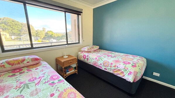 A bedroom with two beds, a small bedside table, a window, and a view of greenery outside. - Northpoint Apartment 5 (SOUTH WEST ROCKS)