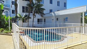 A pool with a white fence, a building with multiple windows, and palm trees. - Ocean Shores Apartment 15 - Bring your own linen (SOUTH WEST ROCKS)