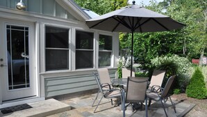Un patio techado con mesa y sillas, un paraguas grande y una casa con ventanas amplias.
