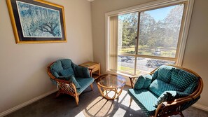 A room with two wicker chairs, a small table, and a large window with a view of a park.