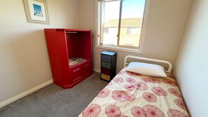 A bedroom with a bed, a red wardrobe, a small desk, and a window with a view of a house.