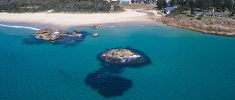 Vista aérea de una zona costera con playa, un promontorio rocoso en el agua y una área residencial con casas y una carretera.