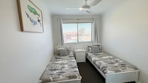 Ein kleines, sauberes Zimmer mit zwei Einzelbetten, einem Deckenventilator, einem Fenster mit Jalousien und einem gerahmten Bild an der Wand.