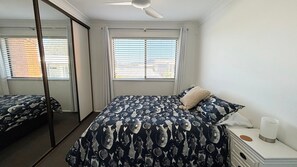 A bedroom with a bed, a bedside table, a mirror, and a window with blinds.