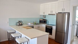 Eine moderne Küche mit weißen Schränken, einem Edelstahlkühlschrank, einer Mikrowelle und einem Toaster. Auf der Arbeitsfläche stehen drei Barhocker und ein Schneidebrett.