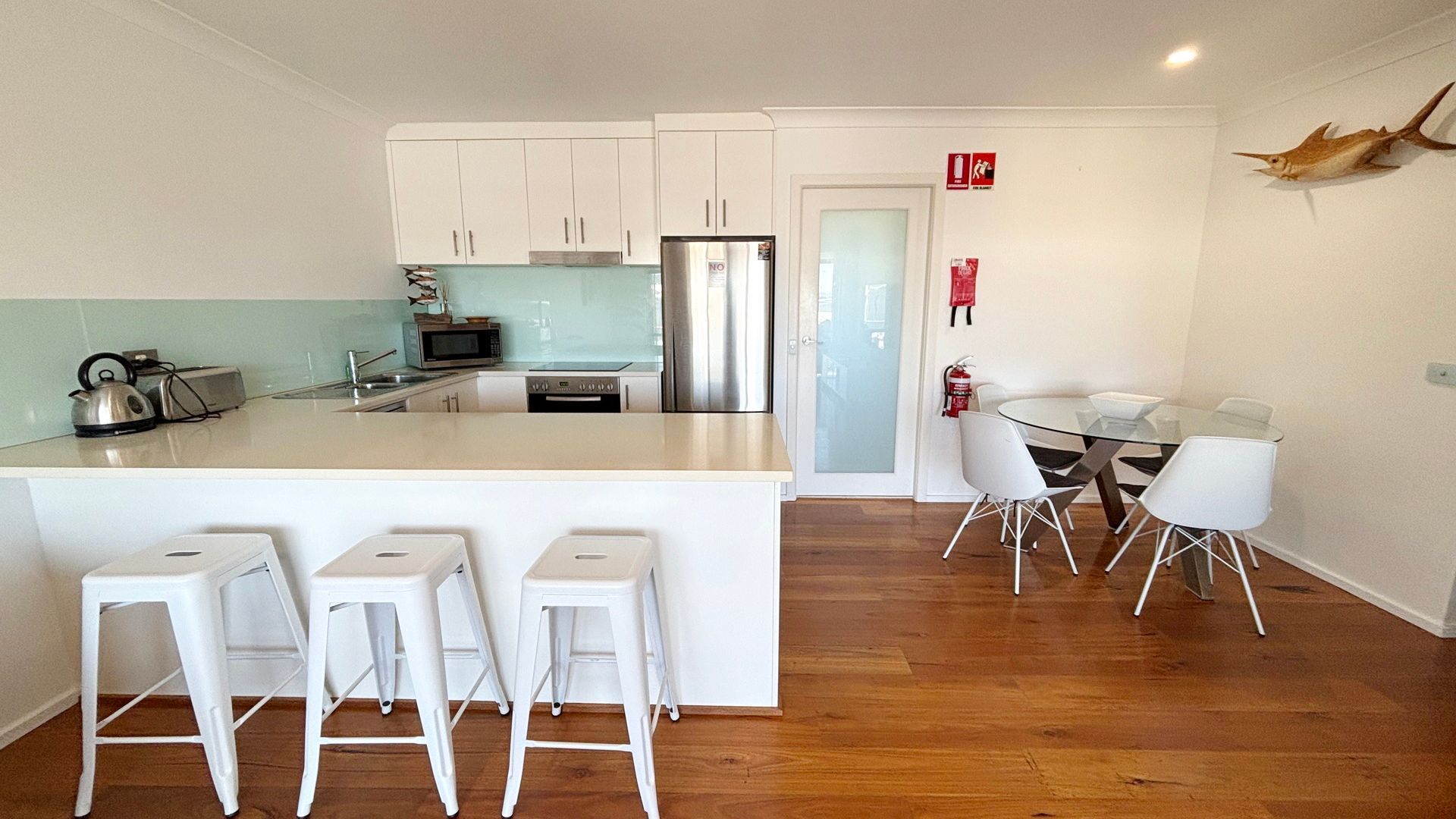 A modern kitchen with white cabinets, a stainless steel refrigerator, and a glass dining table with white chairs.