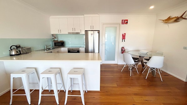 A modern kitchen with white cabinets, a stainless steel refrigerator, and a glass dining table with white chairs. - Seacrest Unit 4 - walk to the beach (South West Rocks)