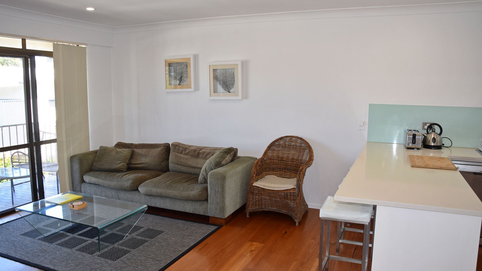 Un salon moderne avec un canapé gris, un fauteuil en osier, une table basse en verre et un coin cuisine équipé d’une bouilloire et d’un grille-pain.