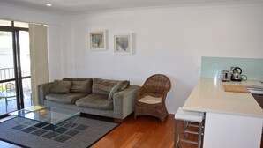 Ein modernes Wohnzimmer mit grauem Sofa, einem Korbstuhl, einem Glastisch und einer Küchenzeile mit Wasserkocher und Toaster.
