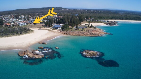 An aerial view of a coastal area with a beach, clear turquoise waters, and rocky outcrops. There are buildings visible in the background, suggesting a residential or resort area. - Pacific Court 1 - walk to the beach - Bring your own linen (South West Rocks)