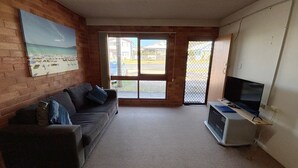 Ein möbliertes Zimmer mit einer Couch, einem Fernseher auf einem Schrank, einem Bild an der Wand und einem großen Fenster mit Blick auf Gebäude und einen Zaun.