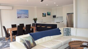 Una sala de estar moderna con un sofá blanco, una mesa de comedor de madera con sillas, una zona de cocina con refrigerador y un cuadro de una playa en la pared.