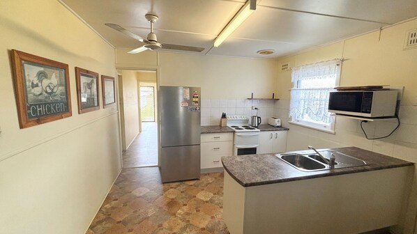 A compact kitchen with a refrigerator, stove, microwave, and sink. There are framed pictures on the wall and a ceiling fan.
