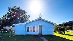 Ein kleines, eingeschossiges blaues Haus mit einer Haustür und Fenstern, umgeben von einem eingezäunten Garten und Bäumen.