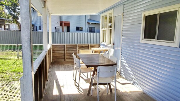 Eine überdachte Terrasse mit Holztisch und weißen Stühlen, eine Holzterrasse und ein Blick auf ein blaues Haus im Hintergrund.