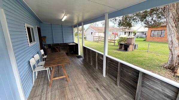 Une terrasse couverte avec une table et des chaises en bois, un petit barbecue et une vue sur une cour arrière avec une clôture et un arbre.