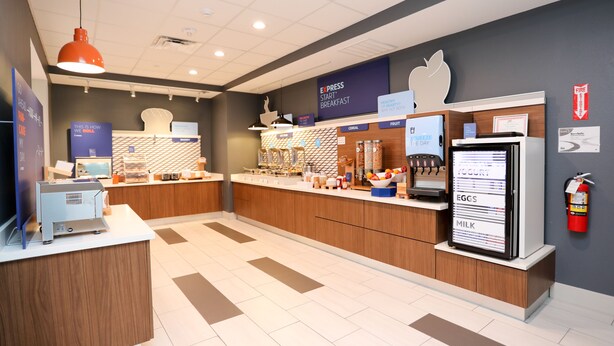 Un espace moderne dédié au petit-déjeuner, doté d’un écran numérique affichant le menu, d’un présentoir à fruits et d’un extincteur fixé au mur.