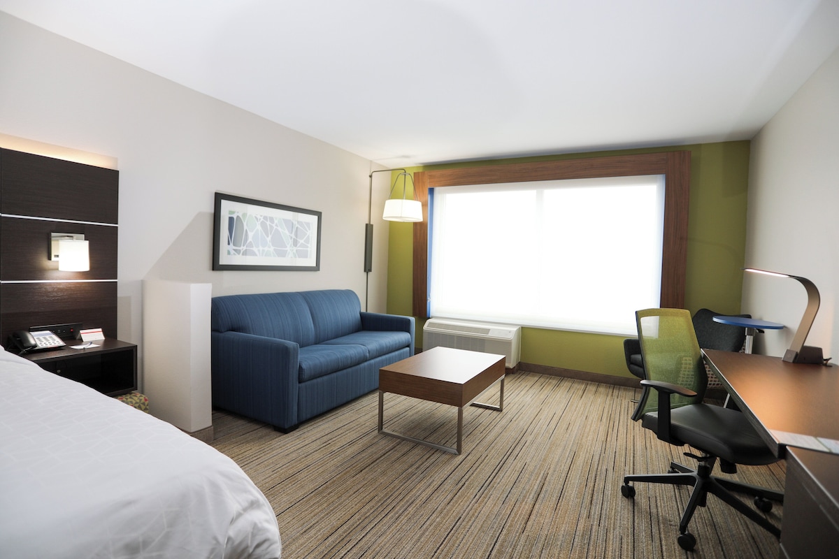A hotel room with a bed, a sofa, a desk with a chair, a small table, a lamp, a picture on the wall, and a window with curtains.