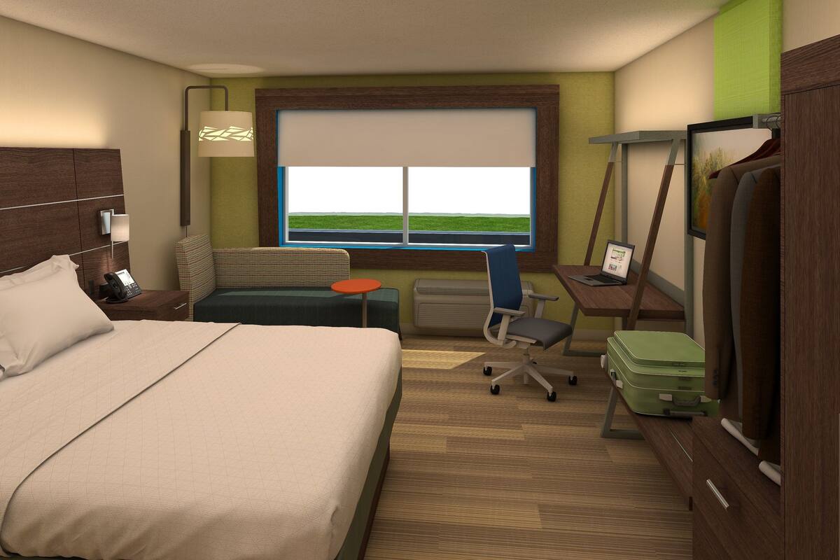 A hotel room with a bed, a desk with a laptop, a chair, a suitcase, and a window with a view of the sky.