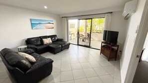 A living room with a black leather sofa, a television on a wooden stand, and a sliding glass door leading to an outdoor area. - Jubilee Apartment 03 (South West Rocks)