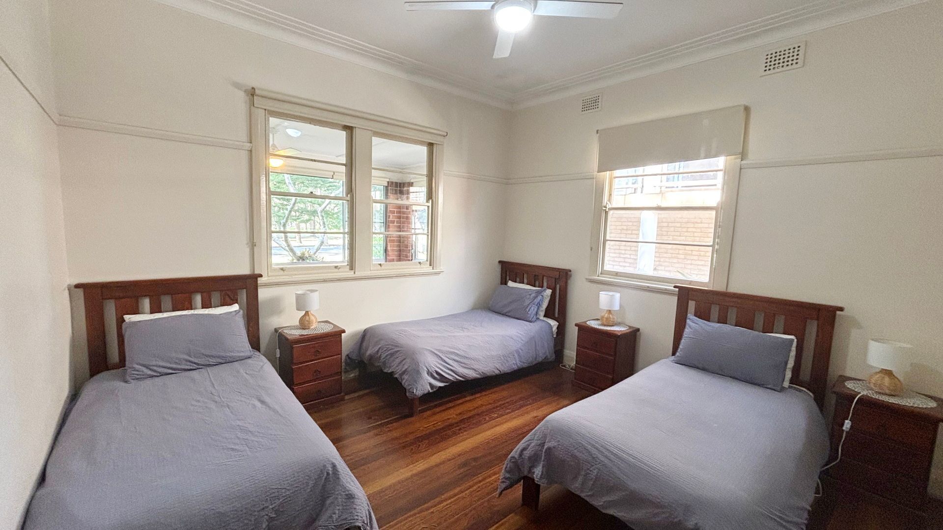 A room with three single beds, each with a wooden headboard, a bedside table, and a lamp. There are windows with blinds, and the room has a wooden floor.