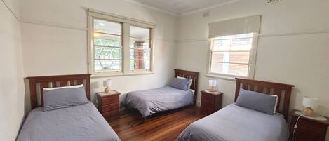 A room with three single beds, each with a wooden headboard, a bedside table, and a lamp. There are windows with blinds, and the room has a wooden floor.