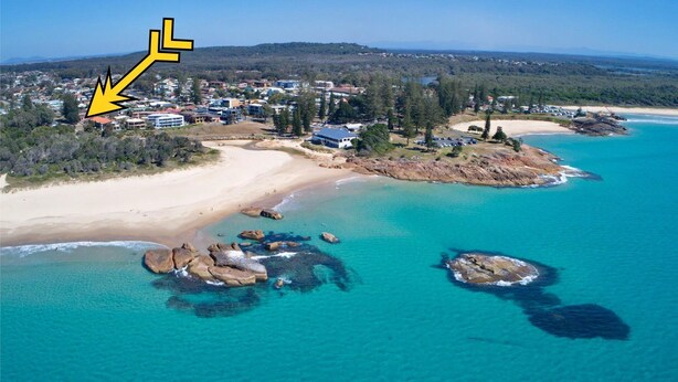 An aerial view of a coastal area with a beach, clear turquoise waters, and a residential area with houses and a large building marked by an arrow. - Yia Yia's Place - walk to the beach - Bring your own linen (SOUTH WEST ROCKS)
