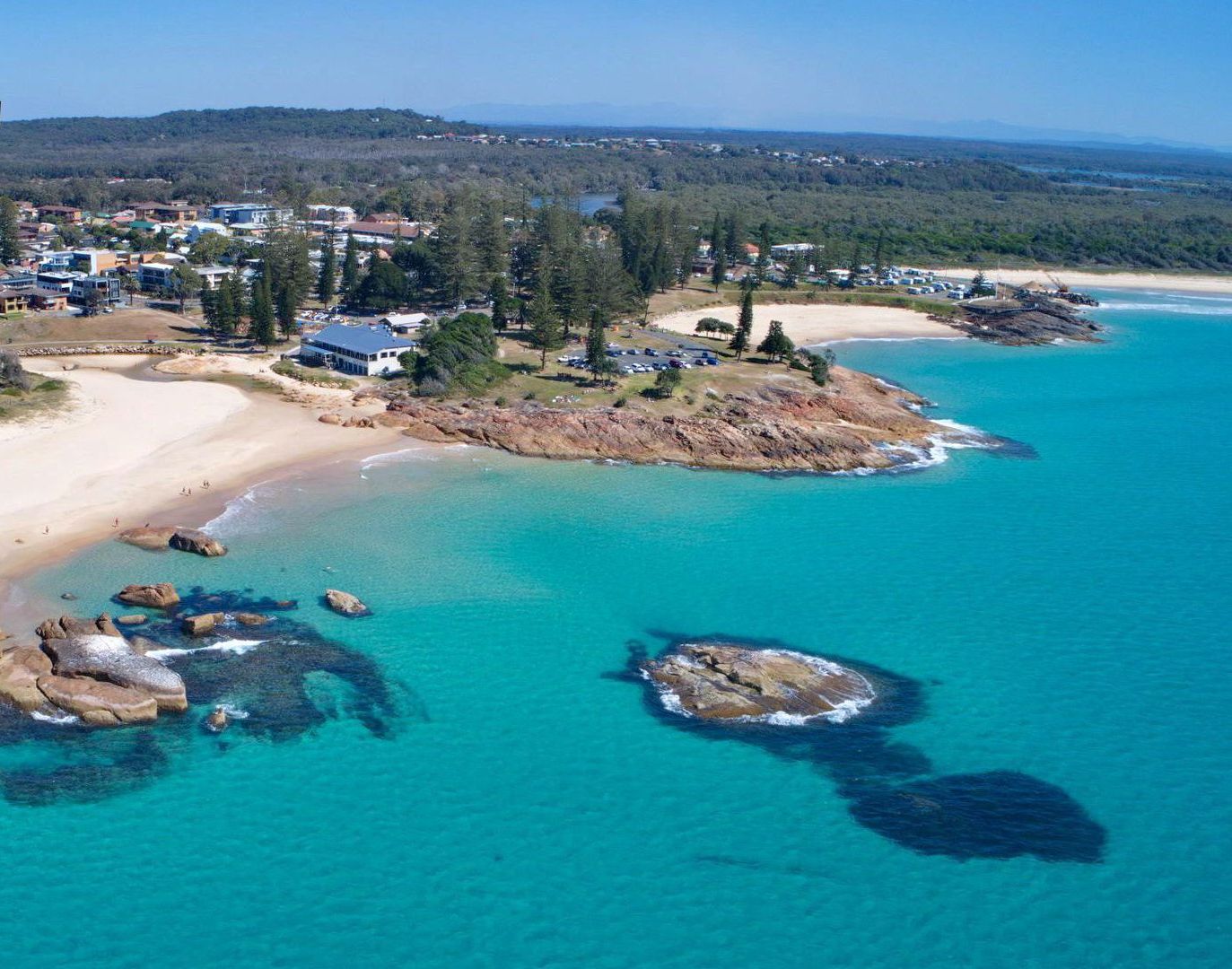 An aerial view of a coastal area with a beach, clear turquoise waters, and a residential area with houses and a large building marked by an arrow.
