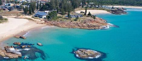 An aerial view of a coastal area with a beach, clear turquoise waters, and a residential area with houses and a large building marked by an arrow.