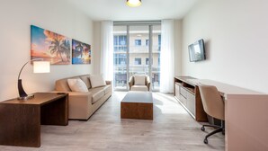 A modern living room with a sofa, coffee table, and desk. There are large windows with a view of a building. - Private Luxury Condos at Costa Hollywood (Hollywood)