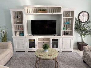 A living room with a television on a white entertainment unit, a round coffee table with a plant, and a patterned rug.