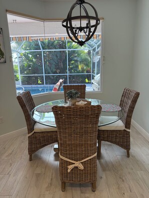 A dining area with wicker chairs and a glass table, a chandelier, and a view of greenery through large windows.