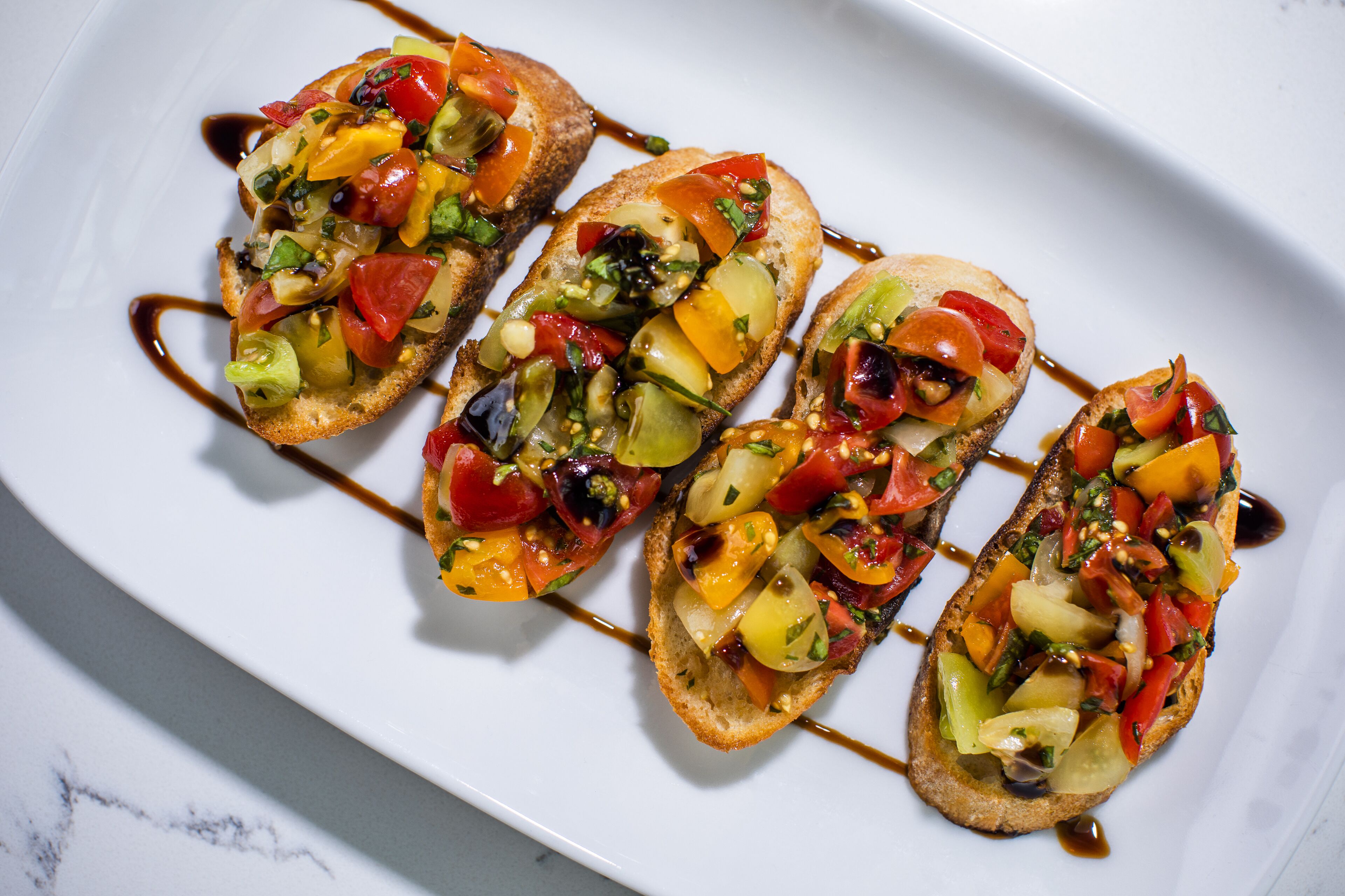 A white plate with four pieces of bruschetta topped with colorful vegetables and a drizzle of sauce.