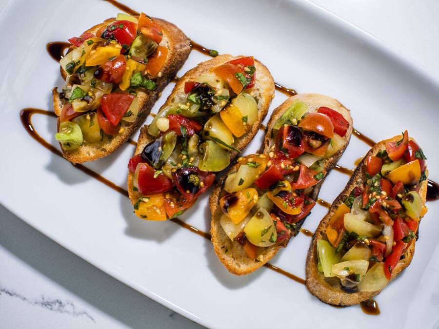 A white plate with four pieces of bruschetta topped with colorful vegetables and a drizzle of sauce.