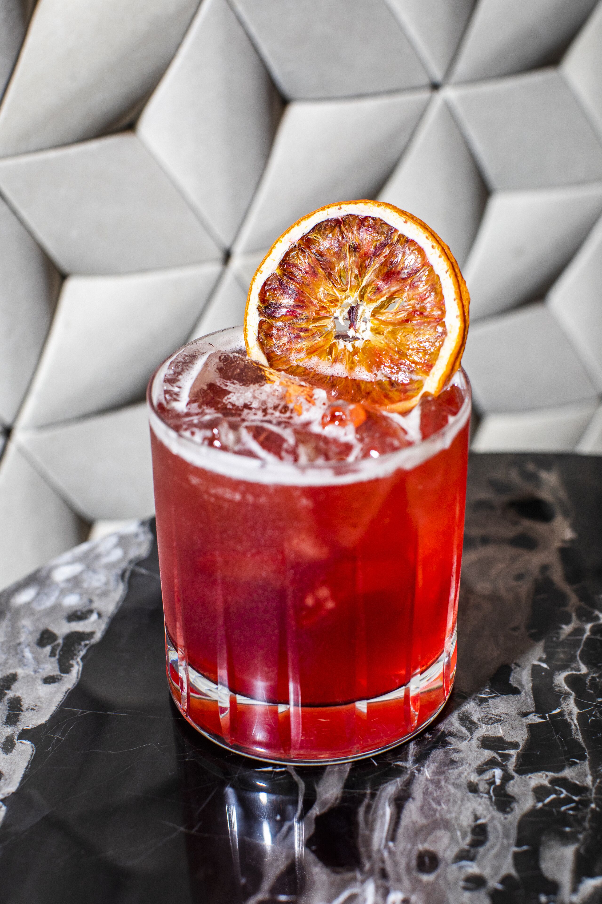 Ein Glas rotes Getränk mit Eiswürfeln und einer Orangenscheibe als Dekoration auf einer spiegelnden Oberfläche.