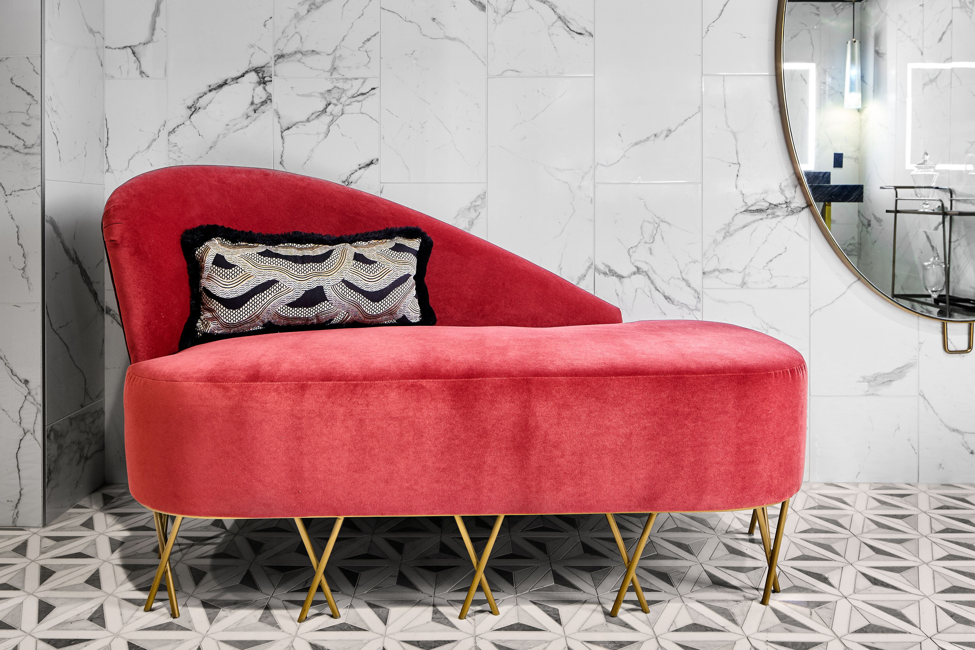 Een rode velourse fauteuil met een gestreept kussen tegen een marmeren muur, een vloer met een geometrisch patroon, en een ronde spiegel met een gouden lijst.