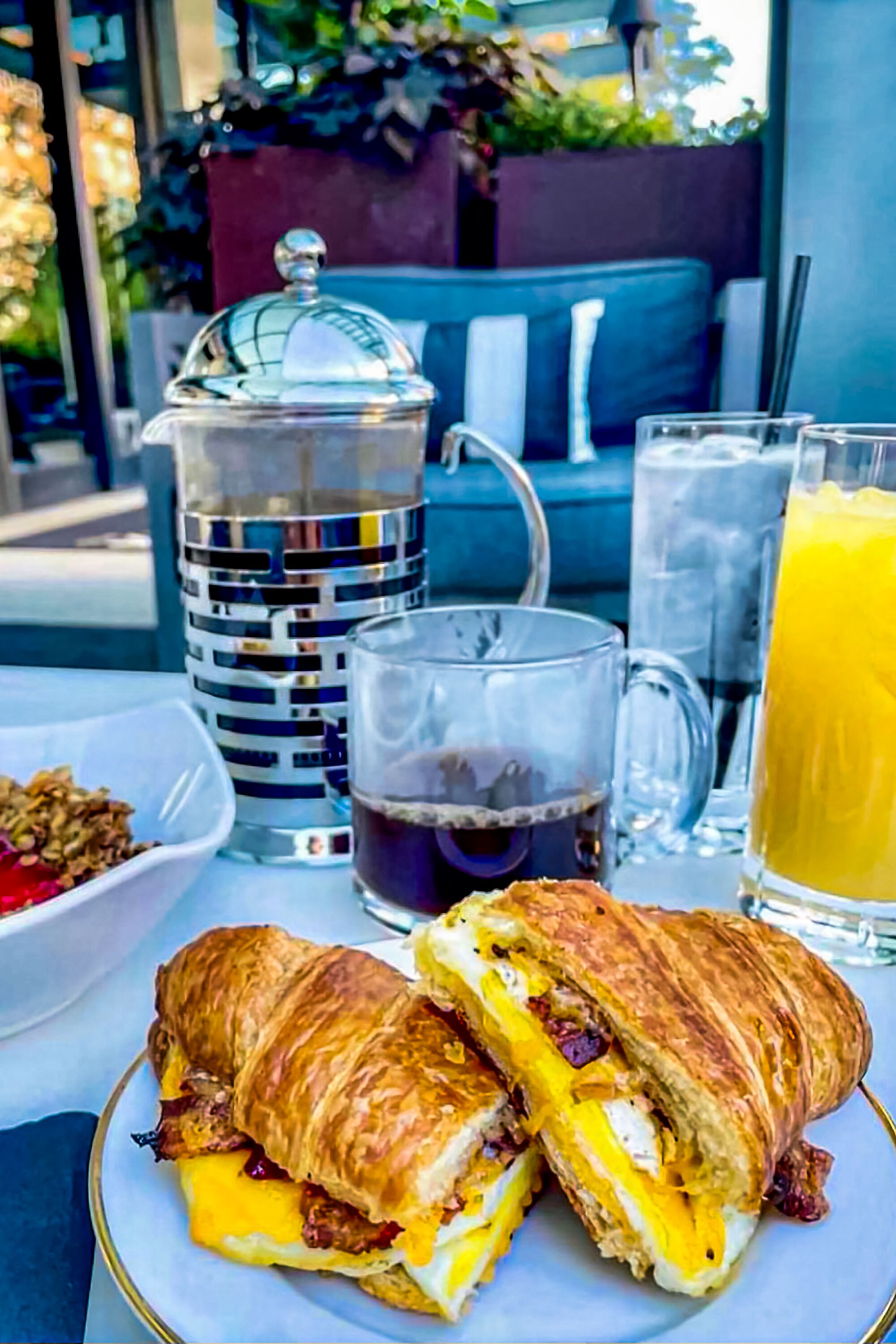 A breakfast setting with a croissant sandwich, a coffee mug, a glass of orange juice, and a pitcher of water.