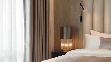A modern hotel room with a large bed, a bedside table with a lamp, and a view of the outside through sheer curtains.