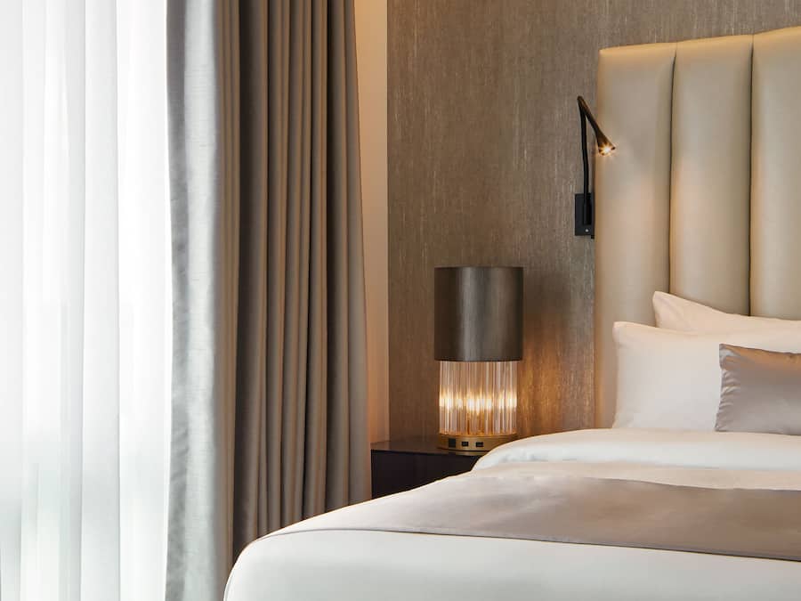 A modern hotel room with a large bed, a bedside table with a lamp, and a view of the outside through sheer curtains.