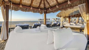 A beachside hotel room with a large bed, multiple lounge chairs, and a thatched roof structure. - Hilton Vacation Club Cabo Azul Los Cabos (San Jose del Cabo)