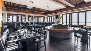 A modern restaurant with a long communal table, individual dining tables, and a central bar area. - Hilton Vacation Club Cabo Azul Los Cabos (San Jose del Cabo)