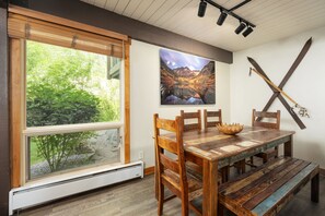 A dining area with a wooden table and chairs, a bench, a window with a view of greenery, and a painting of a mountain landscape on the wall.