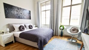 A bedroom with a bed, nightstand, bedside table, window, and a chair. - NEW DESIGN & STYLE Apartment Old Town XXL (Prague)
