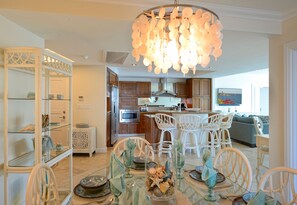 A modern kitchen with wooden cabinets, a stainless steel refrigerator, and a built-in oven. There is a dining area with white wicker chairs and a glass table set for a meal.